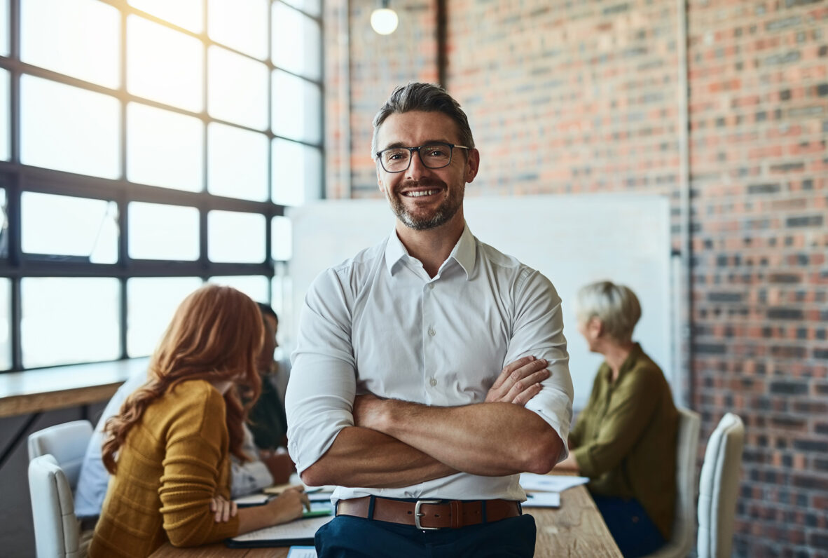 Selbstbewusster Mann in Business-Kleidung steht mit verschränkten Armen vor einem Teammeeting in einem modernen Büro.