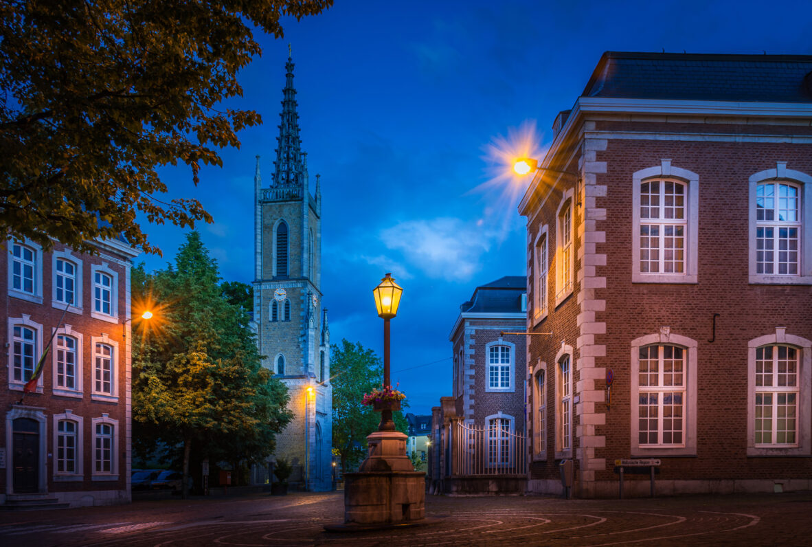 Eupen die alte Post. Historischer Platz in der Abenddämmerung mit beleuchteten Straßenlaternen, umgeben von Backsteingebäuden und einer Kirche mit gotischem Turm.