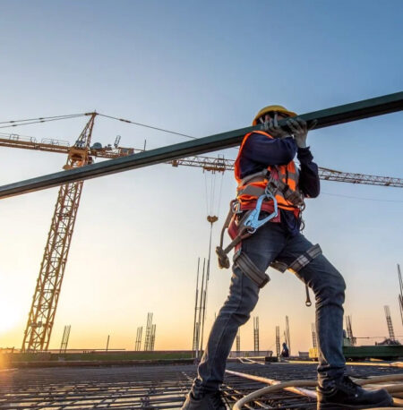 Ein Bauarbeiter mit Schutzhelm, Sicherheitsgurt und Warnweste trägt einen Metallträger auf einer Baustelle. Im Hintergrund stehen Baukräne und Stahlgerüste vor einem Sonnenuntergang.