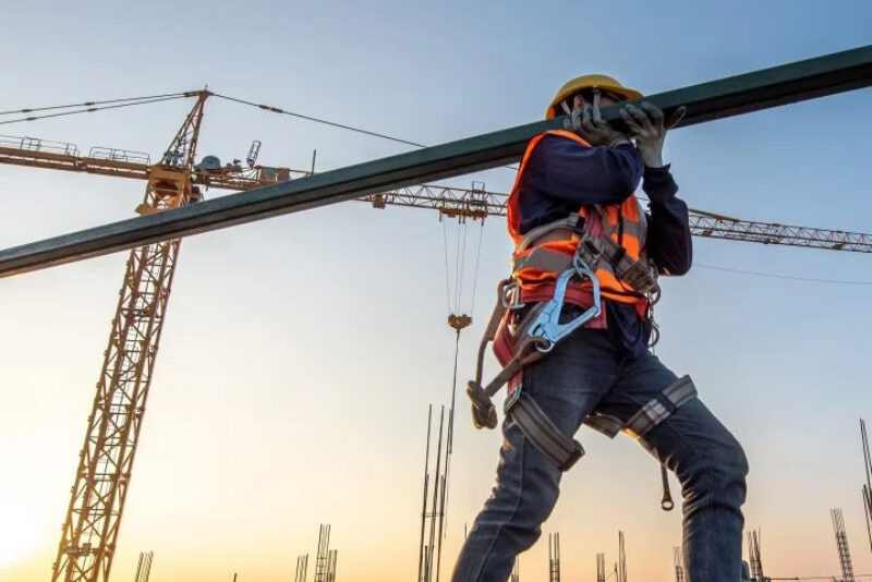 Ein Bauarbeiter mit Schutzhelm, Sicherheitsgurt und Warnweste trägt einen Metallträger auf einer Baustelle. Im Hintergrund stehen Baukräne und Stahlgerüste vor einem Sonnenuntergang.