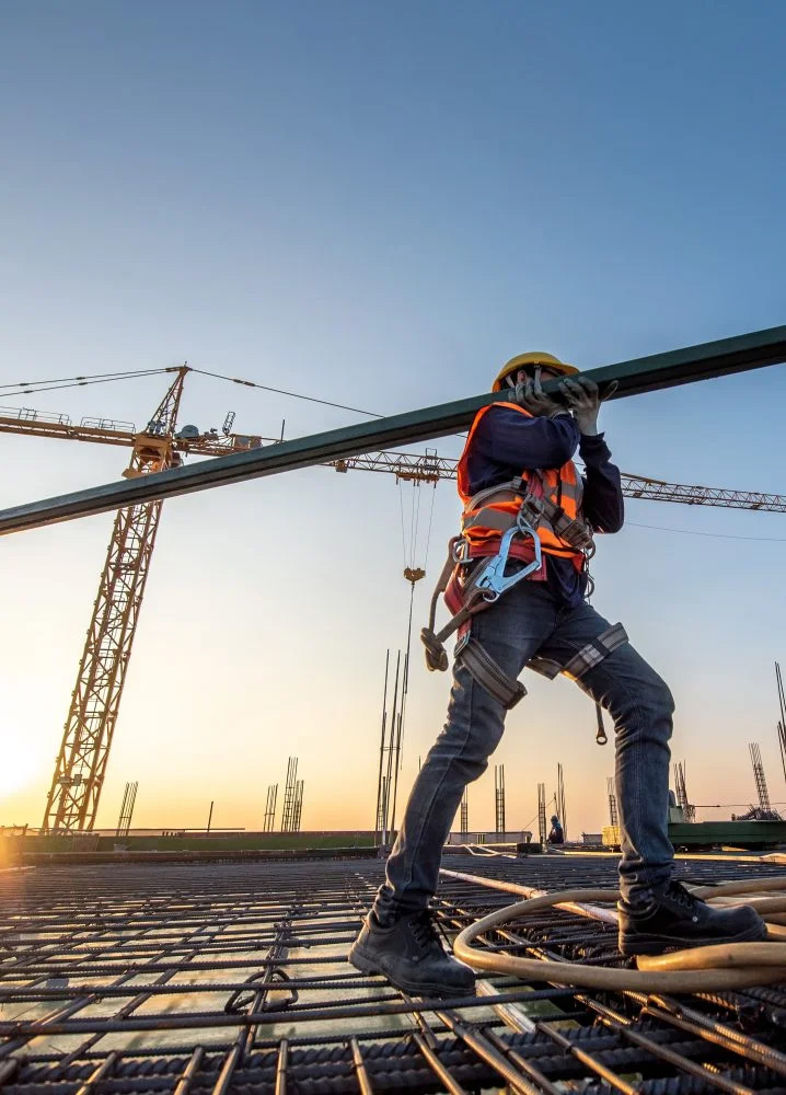 Ein Bauarbeiter mit Schutzhelm, Sicherheitsgurt und Warnweste trägt einen Metallträger auf einer Baustelle. Im Hintergrund stehen Baukräne und Stahlgerüste vor einem Sonnenuntergang.