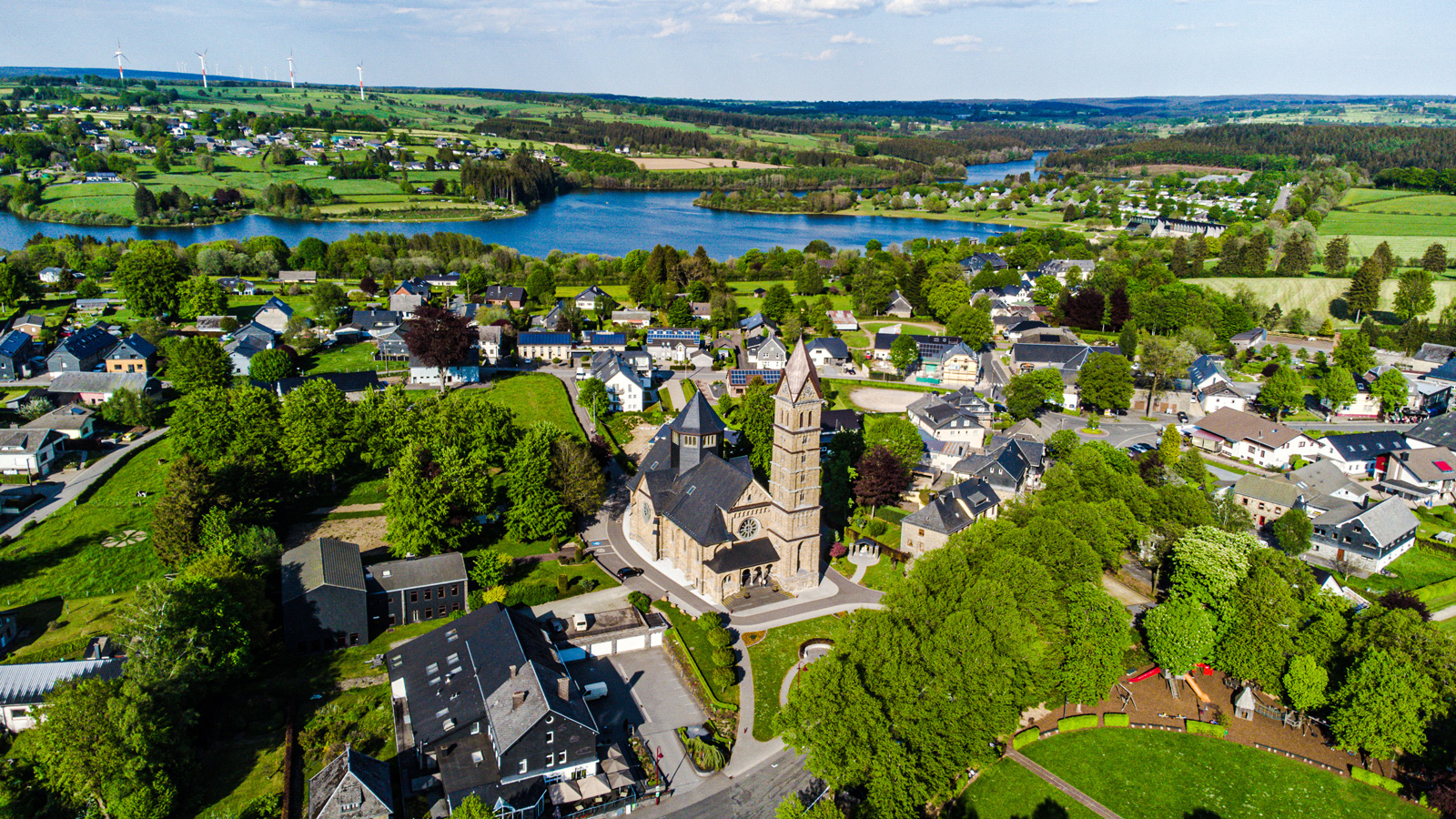 Luftbild von Bütgenbach einer Kleinstadt mit einer großen Kirche im Mittelpunkt, grünen Wiesen, Wohnhäusern und einem See im Hintergrund. Windräder stehen auf den Hügeln in der Ferne.