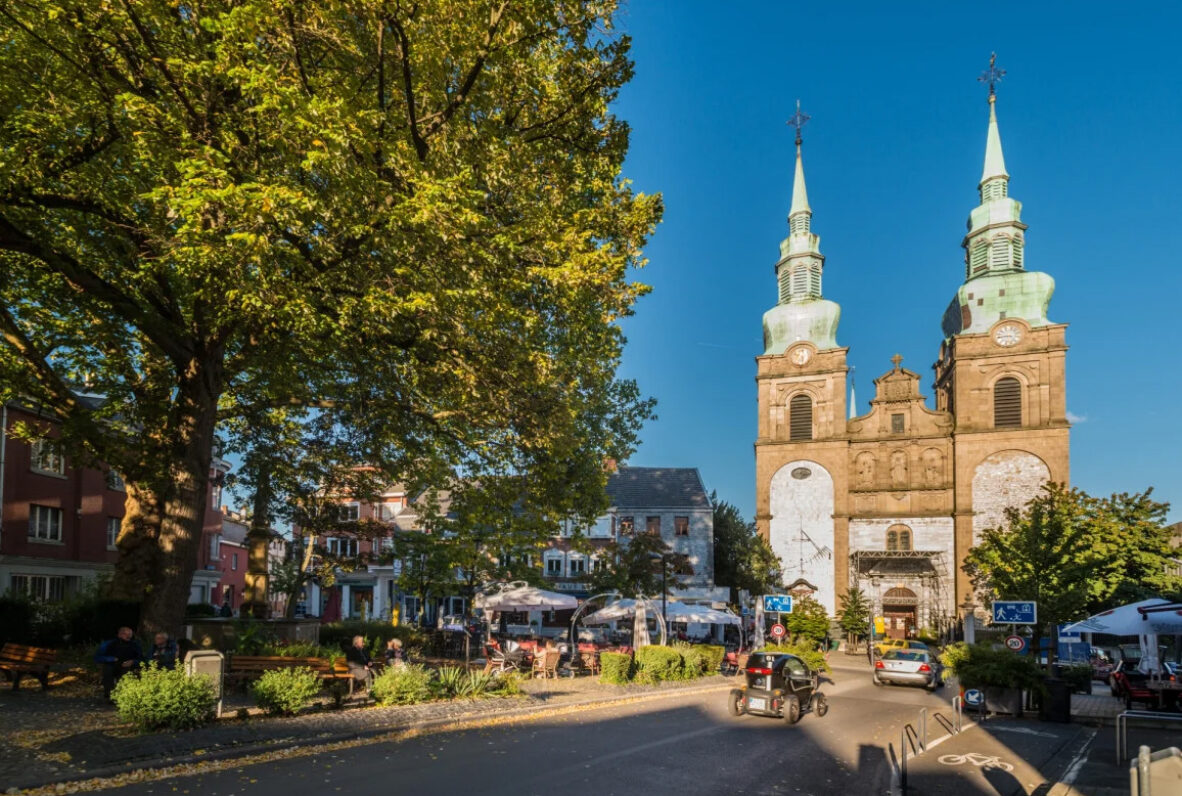 Eupen. Marktplatz mit einer Kirche mit zwei grünen Zwiebeltürmen, umgeben von Cafés, Sitzbänken und Bäumen an einem sonnigen Tag.