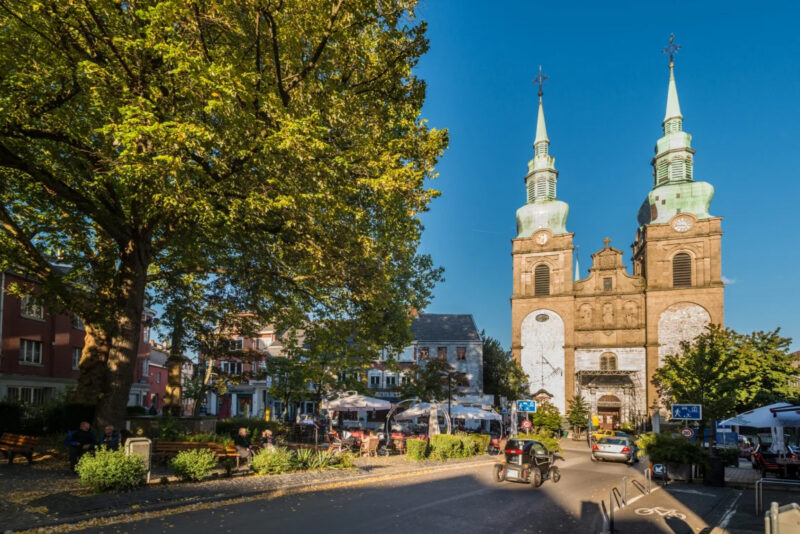 Eupen. Marktplatz mit einer Kirche mit zwei grünen Zwiebeltürmen, umgeben von Cafés, Sitzbänken und Bäumen an einem sonnigen Tag.