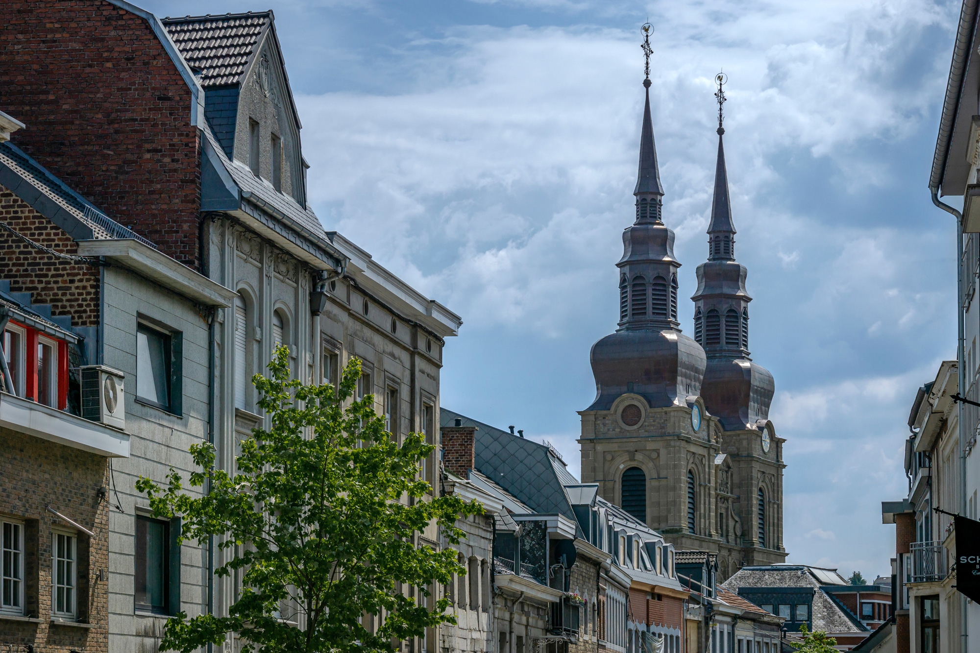 Straßenansicht mit alten Gebäuden in Eupen und einer Kirche mit zwei auffälligen Zwiebeltürmen, die in den Himmel ragen. Der Himmel ist teils bewölkt mit blauen Stellen.