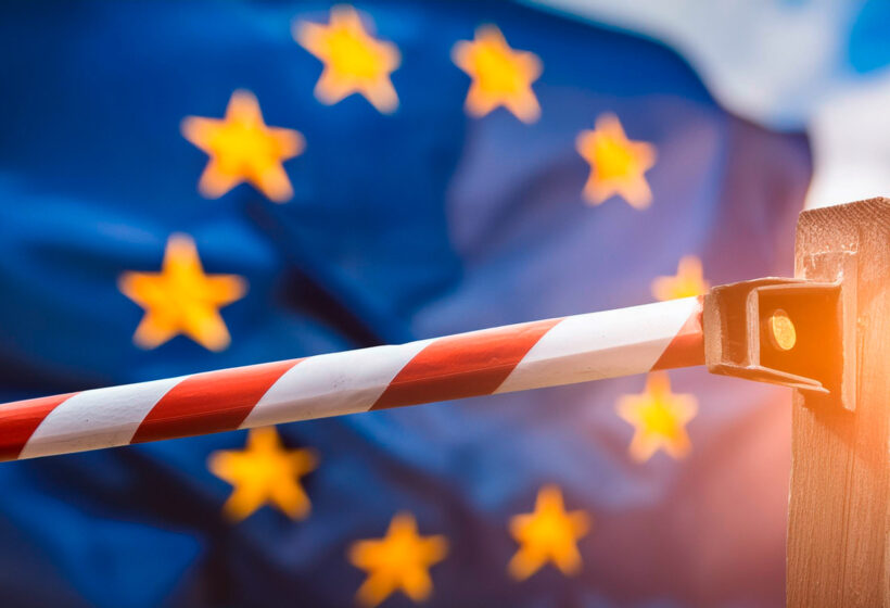 Schranke mit rot-weißen Streifen vor wehender Europaflagge im Hintergrund.