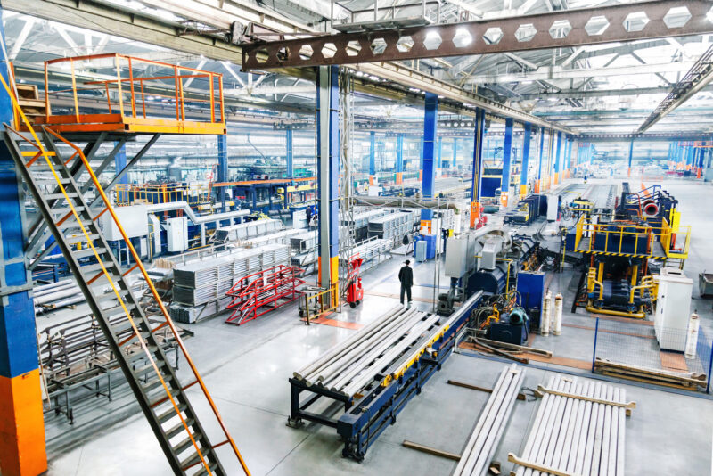 Weitläufige Industriehalle mit Maschinen, Metallteilen und einem Arbeiter, der durch die Fertigungshalle geht.