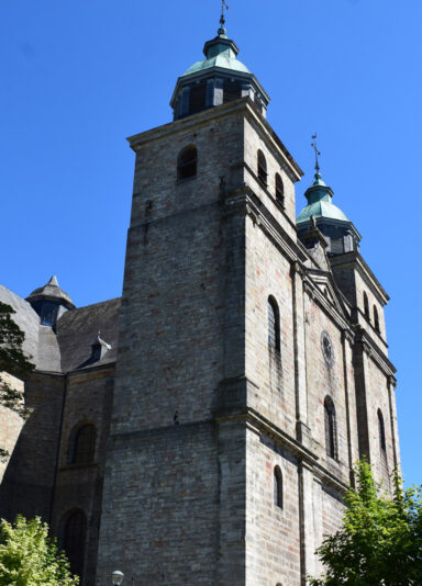 Steinerne Kirche mit zwei hohen Türmen und grünlichen Kupferdächern vor einem klaren, blauen Himmel. Das Gebäude weist historische Architekturdetails auf, und Bäume im Vordergrund sorgen für ein harmonisches Gesamtbild.