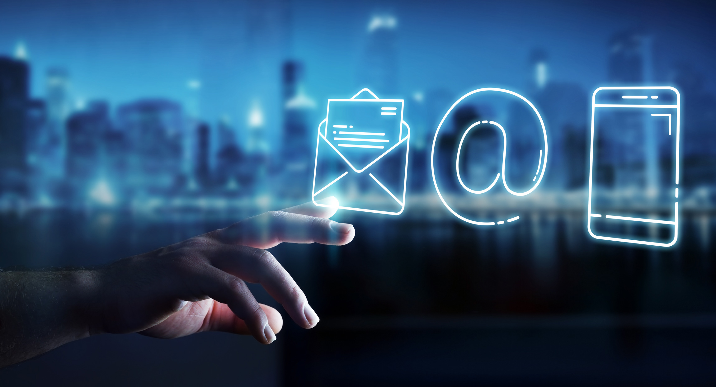 Hand berührt ein digitales E-Mail-Symbol, das zusammen mit einem @-Zeichen und einem Smartphone-Symbol leuchtend vor einer verschwommenen Stadtlandschaft schwebt.
