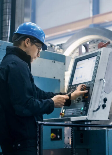 Ein Mann mit blauem Helm und Schutzbrille steht vor einem Maschinenbedienfeld und gibt Daten auf einem Kontrollbildschirm ein. Die Umgebung deutet auf eine industrielle Fertigungshalle hin.