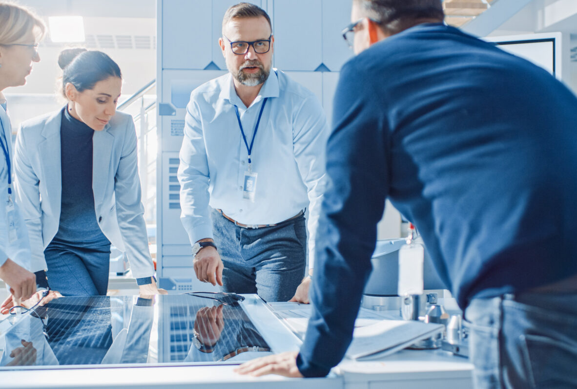 Vier Personen in Business-Kleidung stehen um einen Tisch mit einem technischen Bauteil und besprechen Details in einer modernen, hellen Umgebung.