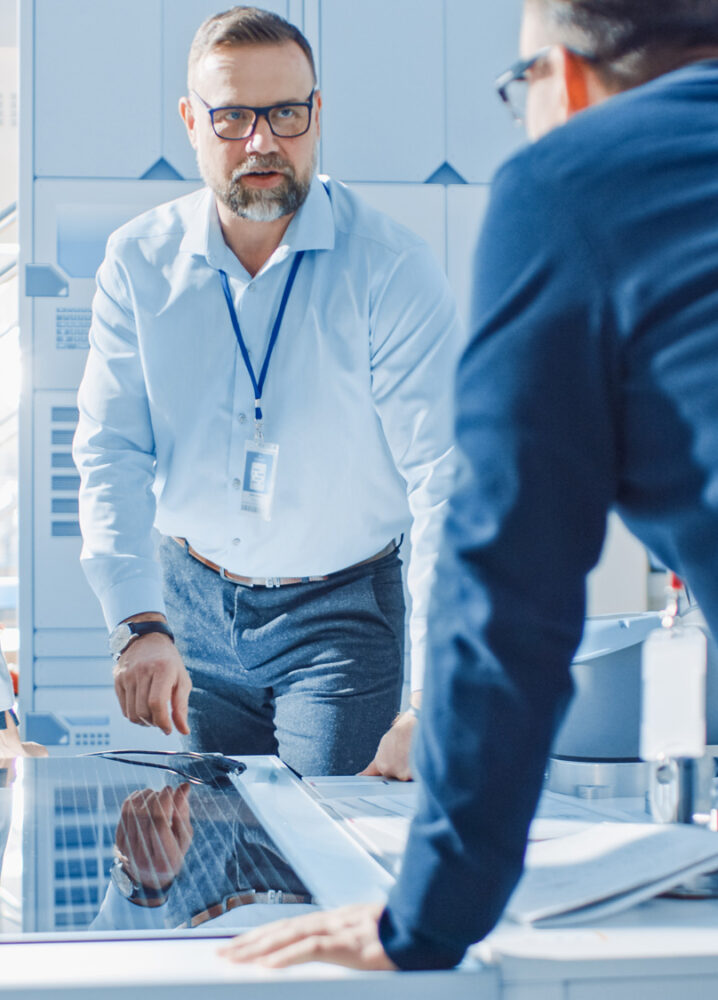 Vier Personen in Business-Kleidung stehen um einen Tisch mit einem technischen Bauteil und besprechen Details in einer modernen, hellen Umgebung.