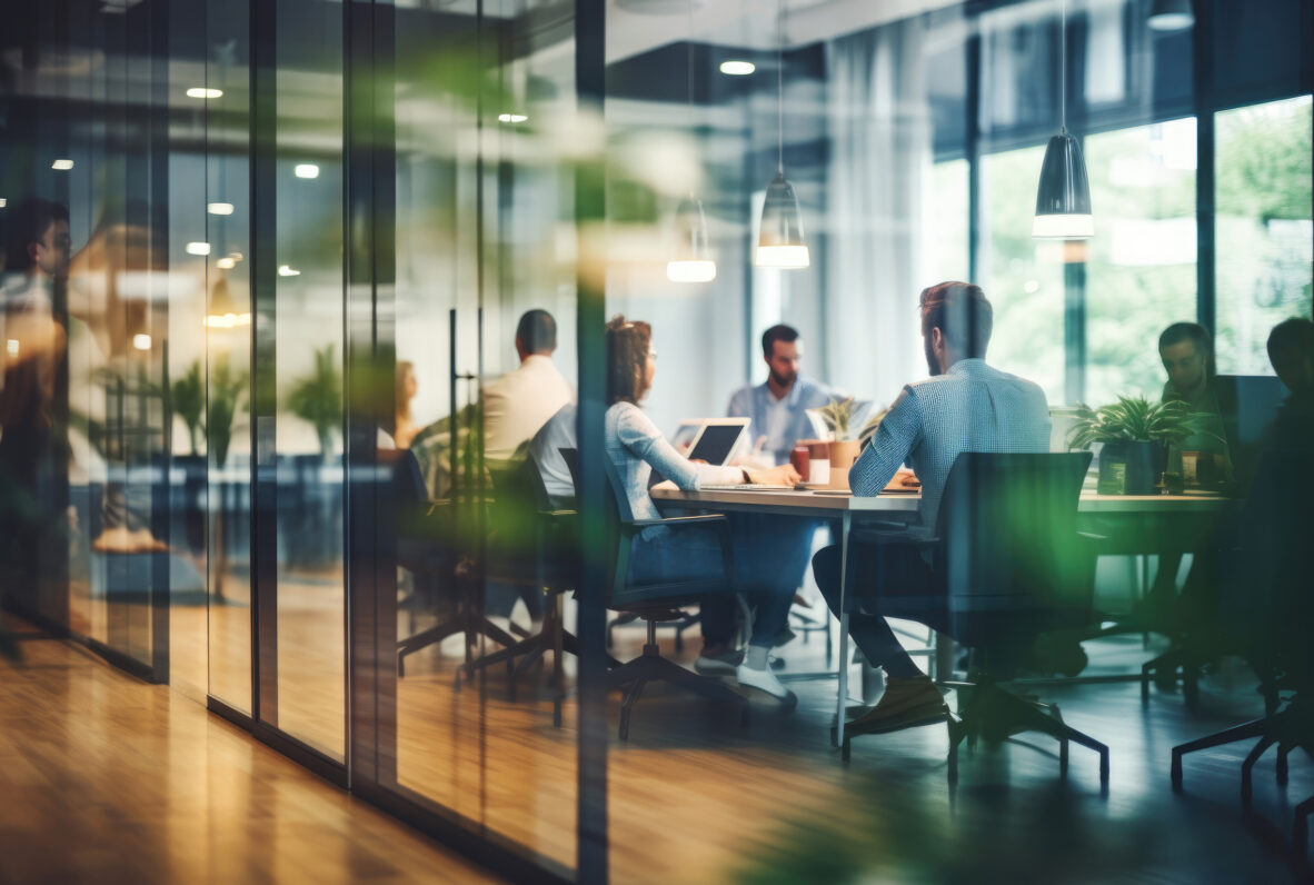 Ein Team sitzt in einem modernen Büro mit Glaswänden und bespricht gemeinsam Projekte. Die Teilnehmer arbeiten mit Laptops und Dokumenten in einer hellen und einladenden Umgebung.