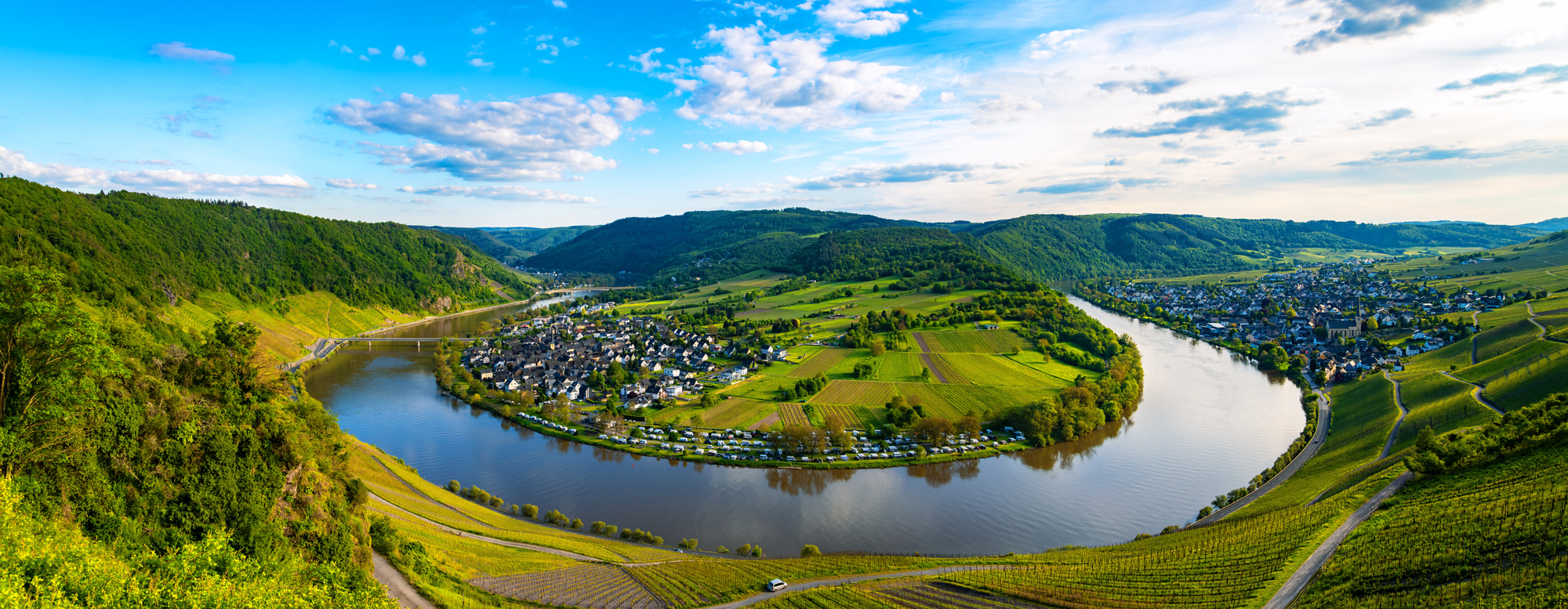 Panoramablick auf eine Flussschleife mit Weinbergen, Dörfern und grünen Hügeln unter blauem Himmel.