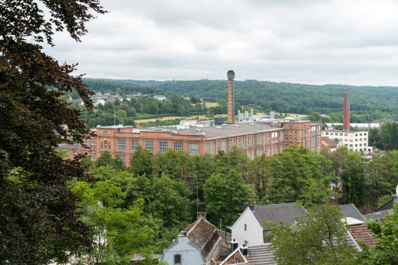 Historische Industrieanlage aus rotem Backstein mit Schornstein, umgeben von Wohnhäusern und grüner Landschaft.