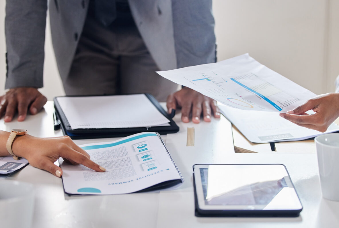 Nahaufnahme eines Besprechungstisches mit Dokumenten, einem Tablet und Kaffeetassen. Mehrere Personen diskutieren und zeigen auf Berichte und Analysen.
