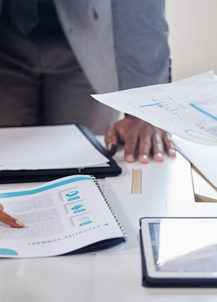 Nahaufnahme eines Besprechungstisches mit Dokumenten, einem Tablet und Kaffeetassen. Mehrere Personen diskutieren und zeigen auf Berichte und Analysen.