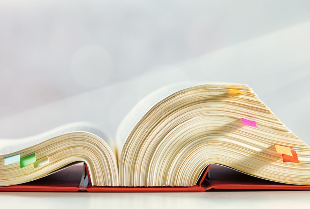 Aufgeschlagenes, dickes Gesetzes Buch mit bunten Markierungen auf einem Tisch, daneben ein Kugelschreiber und ein Smartphone.