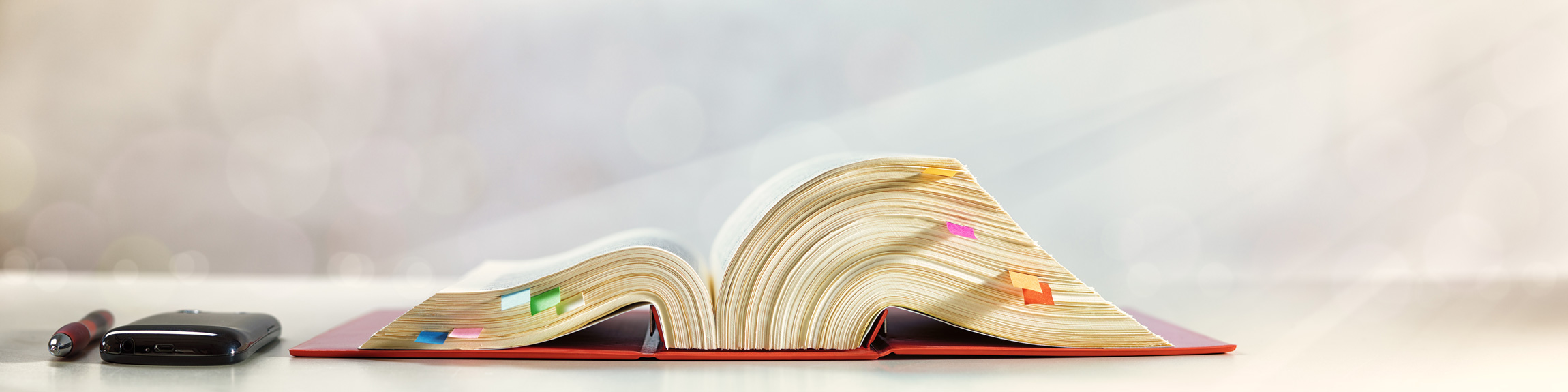 Aufgeschlagenes, dickes Gesetzes Buch mit bunten Markierungen auf einem Tisch, daneben ein Kugelschreiber und ein Smartphone.