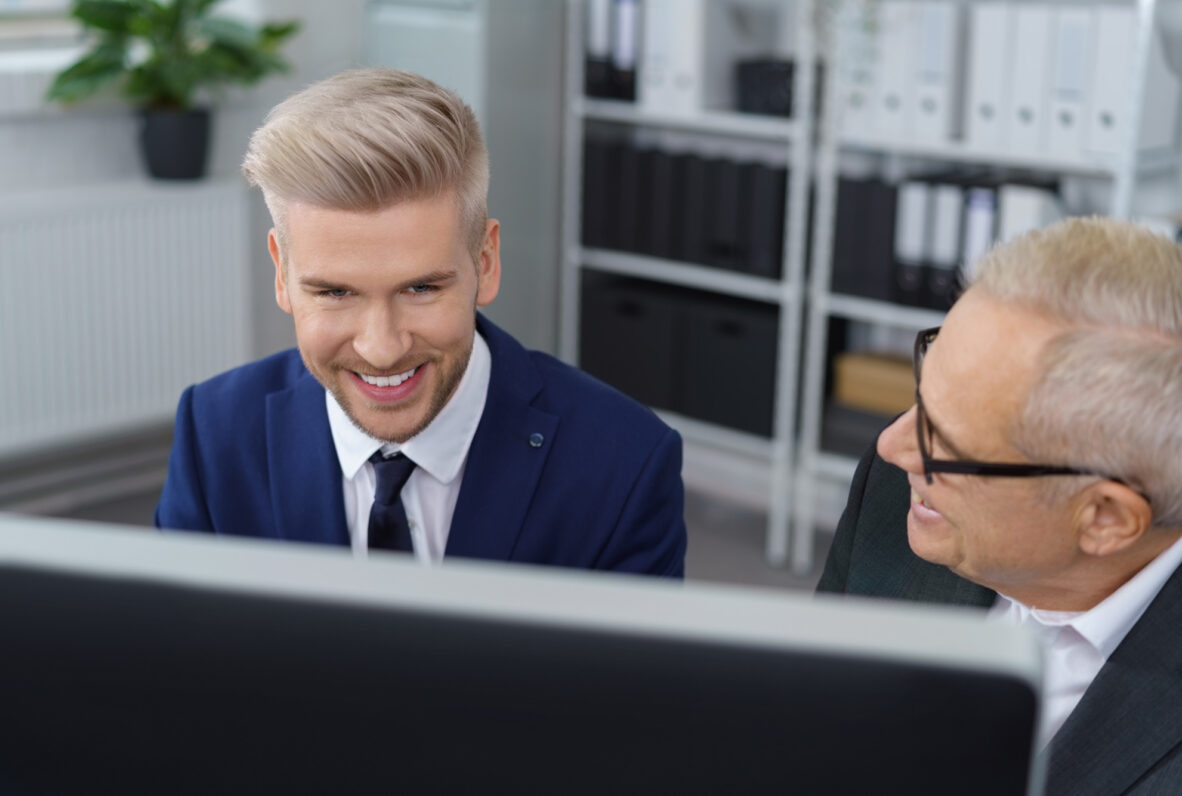 Zwei Männer in einem Büro blicken gemeinsam lächelnd auf einen Computerbildschirm. Der jüngere Mann trägt einen blauen Anzug und hat blondiertes Haar, während der ältere Mann mit grauen Haaren und Brille ihm freundlich zugewandt ist. Im Hintergrund stehen Regale mit Akten und Ordnern.