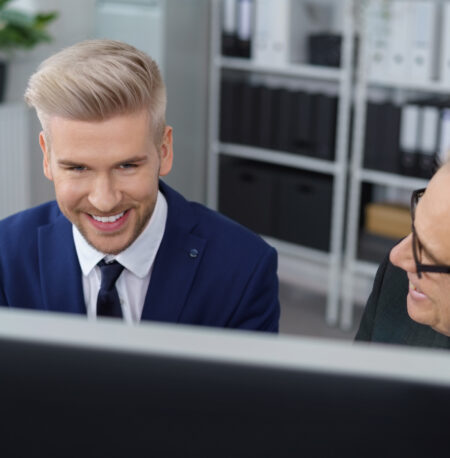 Zwei Männer in einem Büro blicken gemeinsam lächelnd auf einen Computerbildschirm. Der jüngere Mann trägt einen blauen Anzug und hat blondiertes Haar, während der ältere Mann mit grauen Haaren und Brille ihm freundlich zugewandt ist. Im Hintergrund stehen Regale mit Akten und Ordnern.