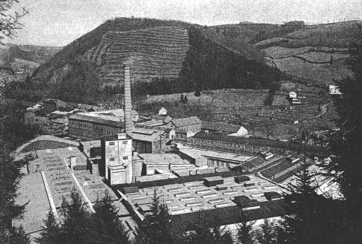 Historisches Schwarz-Weiß-Bild einer Industrieanlage in einer ländlichen Umgebung. Mehrere Fabrikgebäude mit Flachdächern und ein markanter Schornstein dominieren die Szene. Im Hintergrund sind bewaldete Hügel und Felder zu sehen, die das industrielle Zentrum umgeben.